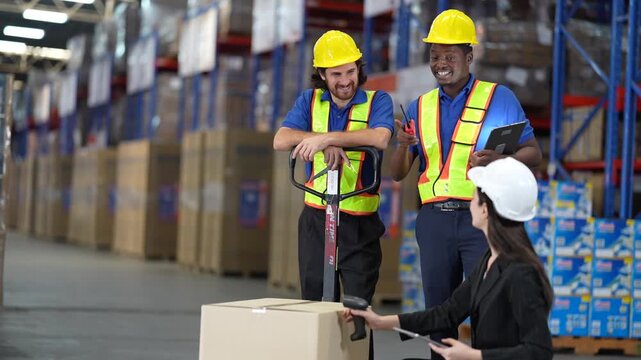 Two warehouse workers with a hand pallet truck smile as a female manager in a white hard hat crouches to check a carton on it