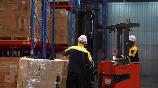 A warehouse worker seen from behind inspects a shrink wrapped pallet while a forklift operator waits at the controls of a red reach truck