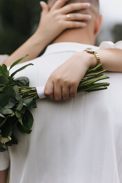 A close-up wedding photo of a bride holding a bouquet and hugging her groom from behind outdoors.