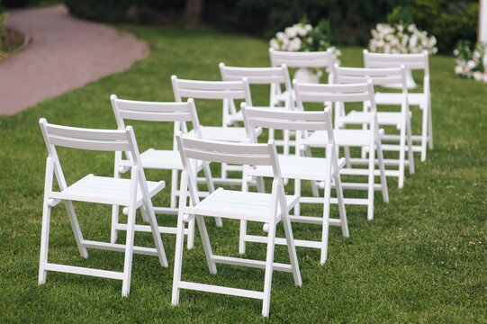 Photo of white wooden chairs on green grass in outdoor park at wedding ceremony.