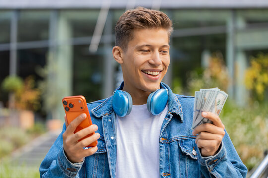 Man holding phone and dollar cash checking amount walking in city street with cheerful smile. Adult guy tourist comparing sum on smartphone counting money feeling pleased during urban stroll outdoors.