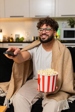 Arabian man at home watches movie on tv holds popcorn laughs comments enjoys calm evening. Egyptian guy on sofa streams show in blanket talks to screen smiles reacts to funny moments at night indoors.