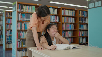 A sleepy young girl yawns widely while studying at a table, while her teacher stands over her attentively inside a library © M Stocker