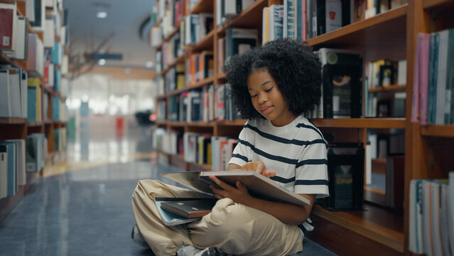 A young girl with curly hair sits cross-legged on a library floor, leaning against a wooden bookshelf while reading a book
