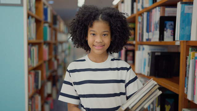 A smiling girl with curly hair and a striped t-shirt stands in a library aisle, carrying a stack of books between wooden bookshelves