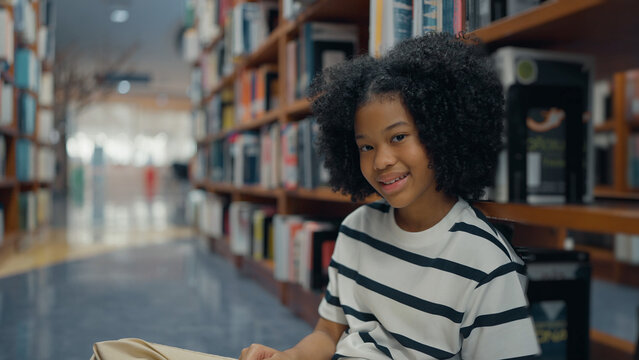 A smiling young girl with a curly afro sits on the library floor, wearing a striped t-shirt and leaning against bookshelves