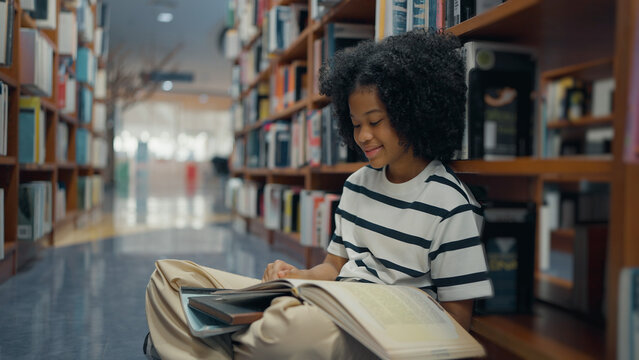 A smiling young girl with a curly afro sits cross-legged on a library floor, leaning against bookshelves while reading a large open book