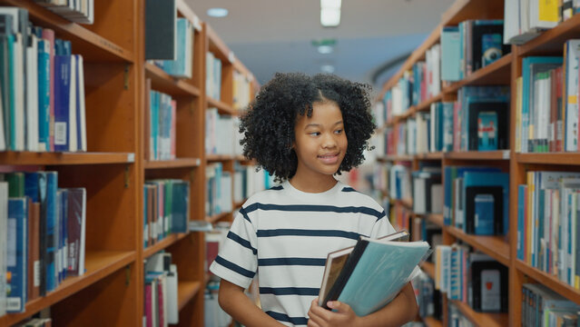 A young girl with curly hair and a striped t-shirt walks down a library aisle, carrying books and folders while looking at the shelves