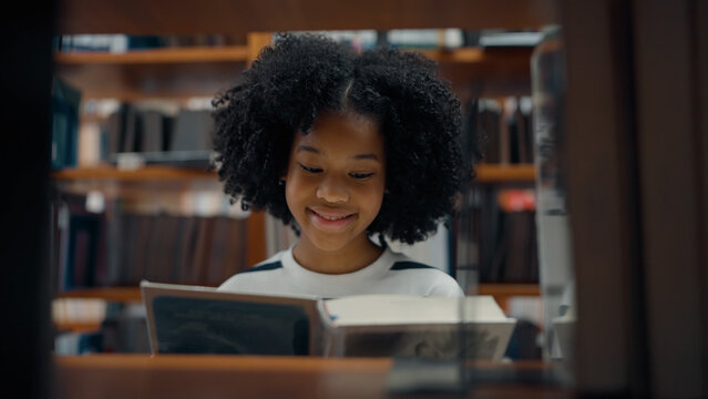A young girl with a curly afro smiles warmly as she reads an open book in a library, framed through a wooden shelf aisle