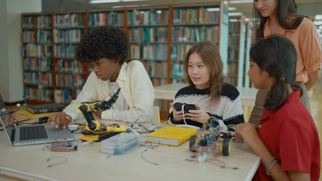 In a library setting, a group of students collaborates on robotics projects, using laptops and remote controls under the guidance of their teacher