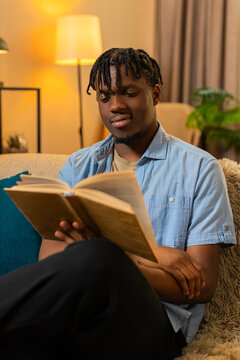 African American young man on home sofa turns pages of a book and finds an interesting idea during reading. Black guy on home sofa pauses to think about a helpful thought and enjoys leisure indoors.
