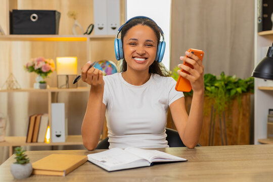 African American young woman on home table takes language test on smartphone and sees right answer. Black girl makes notes in notebook while studying words with headphones in an online lesson at home.