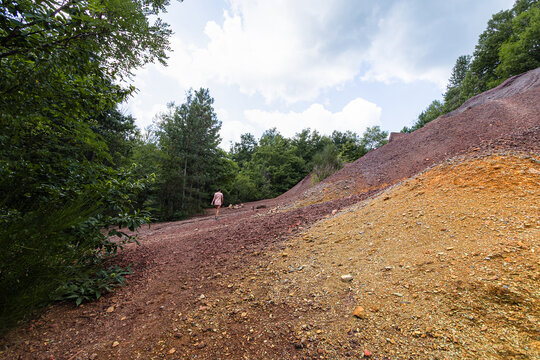 A woman walks along the vibrant red and orange mineral slopes bordered by green trees at Le Roste di Montieri. These surreal environments reveal the deep geological impact of ancient Tuscan mining.