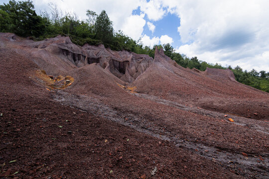 Eroded red mineral hills and dark gravel slopes stretch under a bright cloudy sky at Le Roste di Montieri. These striking badlands reveal the deep geological history and raw industrial heritage.