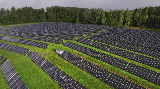 Cinematic aerial drone footage flying over an extensive solar farm with long rows of photovoltaic panels surrounded by green vegetation and forest. Large-scale renewable energy infrastructure captured
