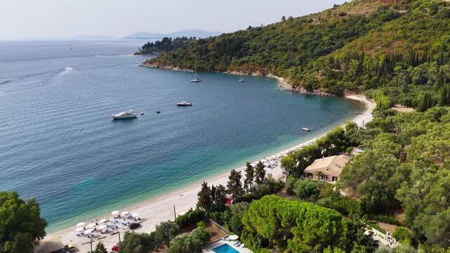 Aerial drone view of Kerasia beach in northeast Corfu, Greece with clear turquoise water, white pebbles, boats and trees