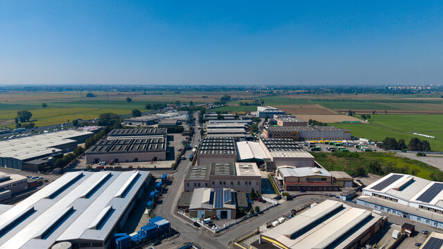 Aerial view of the industrial zone with large warehouses, flat roofs, and surrounding green fields under a clear blue sky in San Giuliano Milanese, Lombardy, Italy.