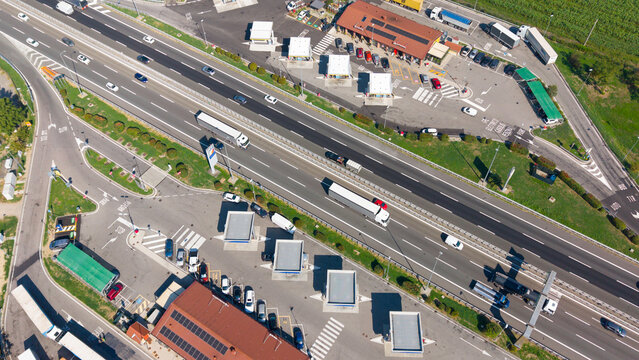 Aerial view of a busy motorway rest stop with parked cars and trucks, adjacent to a highway with flowing traffic in San Giuliano Milanese, Lombardy, Italy.