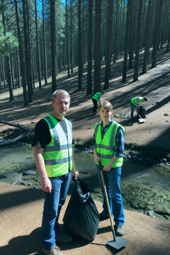 Portrait of Caucasian middle aged man and teenager volunteering in forest, collecting litter near stream with cleanup team. Ideal for environmental campaign, conservation outreach, vertical