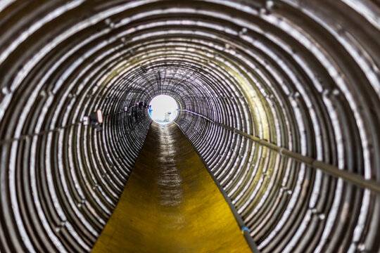 A long corrugated metal tunnel with ribbed concentric rings leads toward a bright circular exit, illuminated by side lights and lined with pipes along the wall