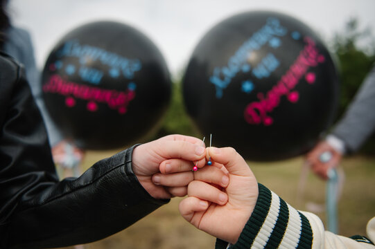 Children hands holding sharp pins to pop black gender reveal balloons for twins outdoors.