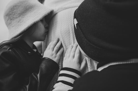Black and white photo. Cute children gently touching and kissing the belly of their pregnant mother outdoors during a family twin pregnancy photoshoot.