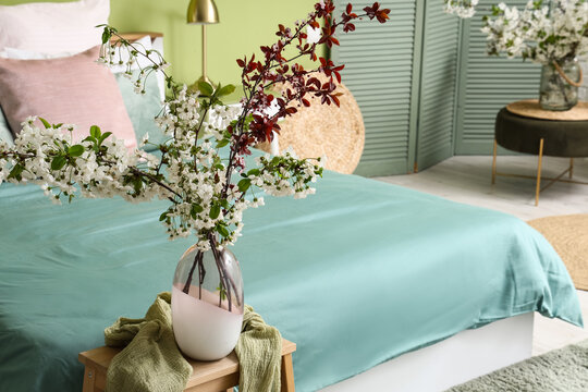 Vase of blooming tree branches on coffee table near bed in stylish bedroom, closeup