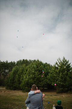 Loving couple embracing while looking up at floating confetti and balloons against the sky during a twin gender reveal celebration in a forest.