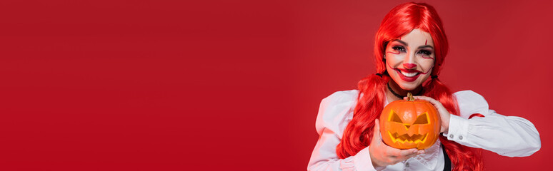 Naklejka premium Young woman with bright red hair and clown makeup smiles while holding a pumpkin, banner 