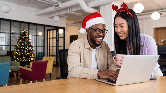 Man and woman wearing Christmas hats work on laptop in modern office decorated for holiday season with Christmas tree in the background
