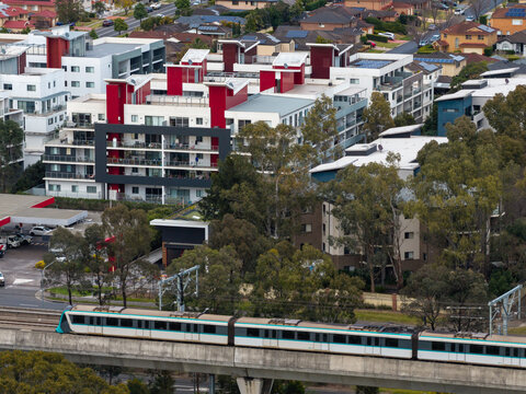 Aerial view of a Sydney Metro train passing by a modern apartment precinct and suburban housing development with lush trees in Kellyville Ridge, New South Wales, Australia.