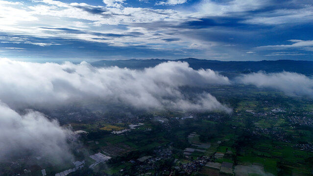 Aerial view of lush green agricultural fields and rural settlements partially obscured by low-hanging clouds under a dramatic sky in Sukabumi Regency, West Java, Indonesia.