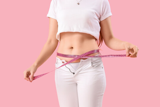 Young woman in tight pants measuring her belly on pink background. Weight gain concept