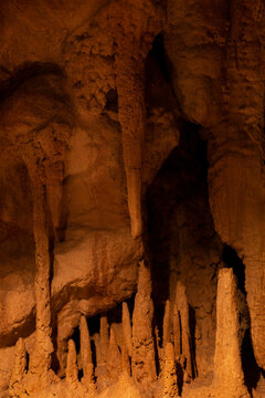 Cave interior showing stalactites and stalagmites formed from mineral deposits, lit by warm amber light that reveals rough rock texture and depth 