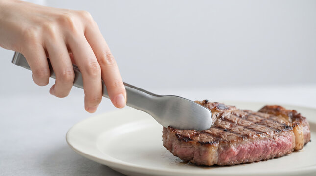 トングで焼きたてのステーキ肉をつかむ手元と料理イメージ