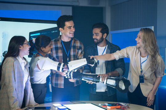 A diverse engineering team gathers in a tech lab to test a robotic arm, smiling and interacting while working on a prototype project during the workday in a modern office interior space.