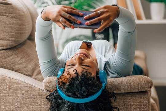 Smiling young woman relaxing in armchair, lying upside down, wearing blue wireless headphones and taking selfie with smartphone, cheerful candid lifestyle