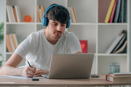 Young Caucasian man wearing headphones, writing notes while studying online with a laptop at a wooden desk, focused and concentrated, home office with bookshelf background