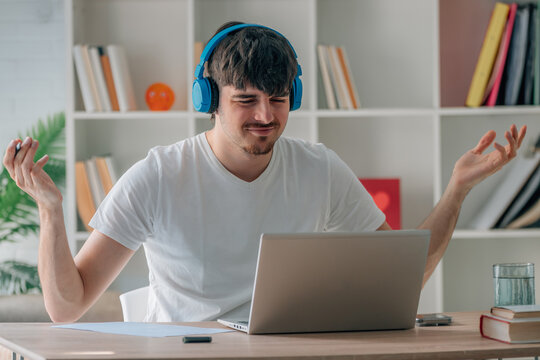 Confused young Caucasian man wearing headphones gesturing with hands while looking at laptop screen during video call, expressing doubt and uncertainty while working from home