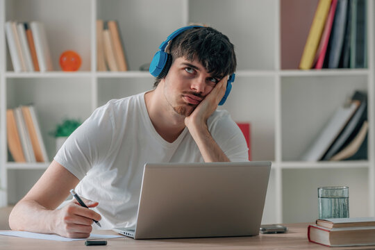 Tired young student man sitting at a desk with a laptop, resting his head on his hand while looking bored and unmotivated, holding a pen over a notebook in a home office