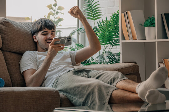 Excited young Caucasian man sitting in armchair celebrating victory while playing game on smartphone, raising fist in success gesture at home