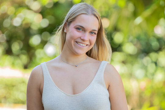 Portrait of a happy young Caucasian woman smiling outdoors, natural sunlight, green blurred background, candid lifestyle photography