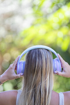 Rear view of a young Caucasian woman outdoors holding purple wireless headphones on her head, enjoying music in a green natural setting