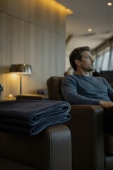 Fototapeta premium A man is comfortably settled in a cozy lounge area, surrounded by a pile of soft blankets. The warm lighting creates a serene atmosphere as he gazes thoughtfully out the window, lost in his own reflec