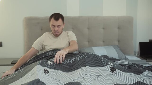 Man sitting up in bed shifting duvet while waking and checking under covers, holding smartphone, with tufted headboard in modern bedroom morning light near nightstand