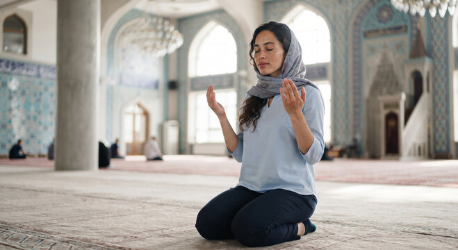 Muslim woman in hijab kneeling and praying on a carpet inside a mosque