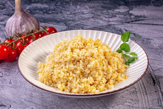 Cooked bulgur wheat in white ceramic bowl with fresh cherry tomatoes, garlic bulb, and green pea shoots on textured gray surface
