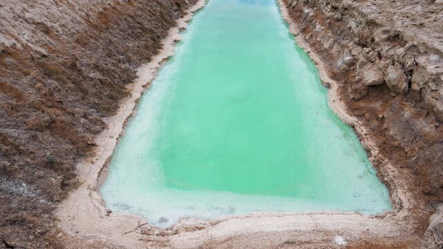 Stunning aerial shot slowly revealing a long, narrow brine channel filled with vibrant turquoise water, carved into the barren, salty landscape of a vast desert under a clear blue sky