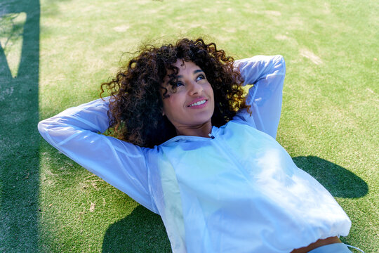Woman with curly hair lying on artificial grass with hands behind head, smiling and looking up, relaxed and enjoying sunny outdoor break