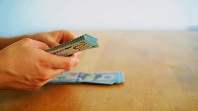 Side view of hands counting stack of 100 US dollar bills over wooden desk. Man counting cash money notes for payment, business salary, savings or financial investment in bright modern office space.